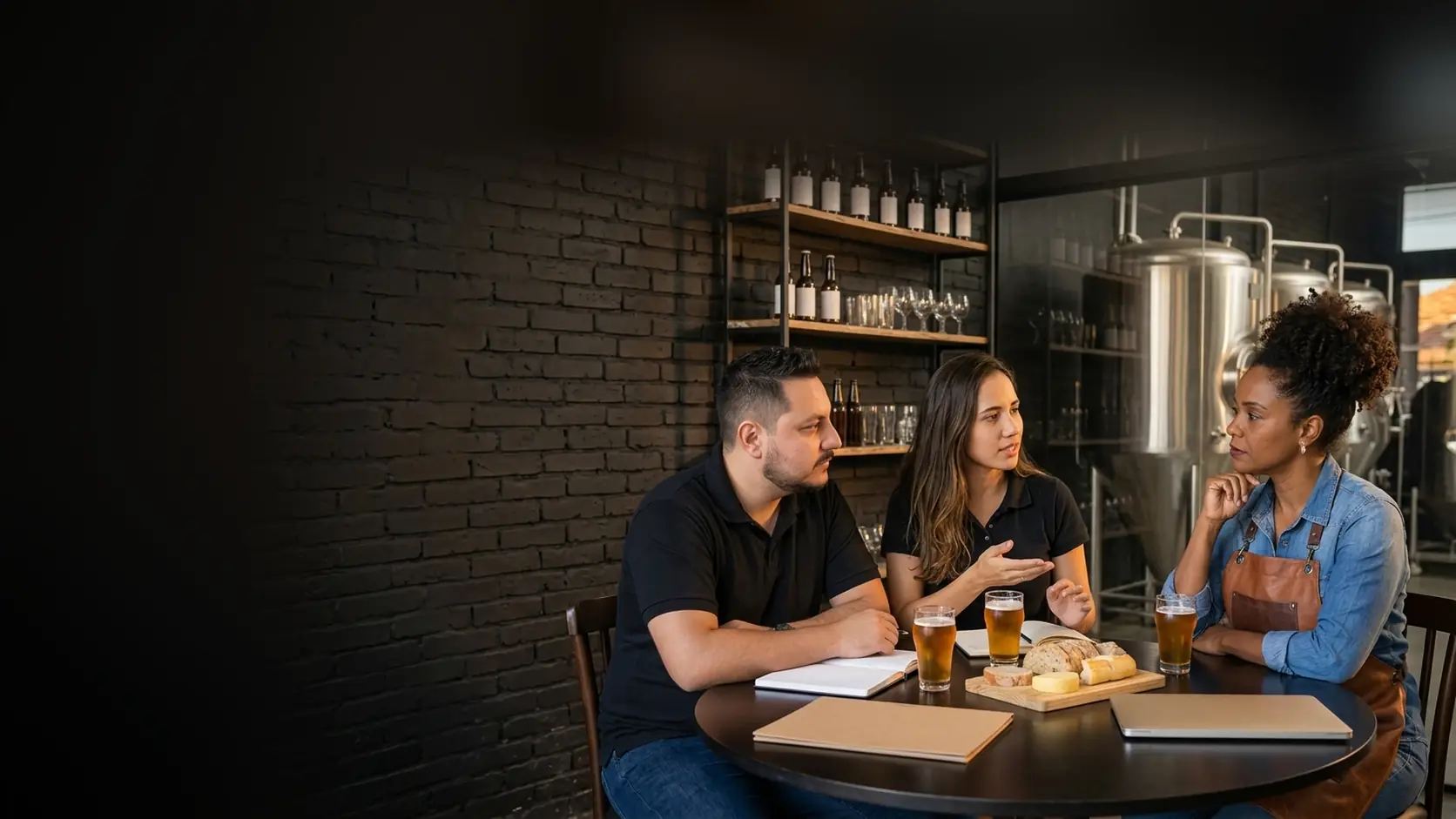 Léo e Lari conversam com a dona de uma cervejaria artesanal em mesa redonda, com copos de cerveja, cadernos e tanques de inox ao fundo