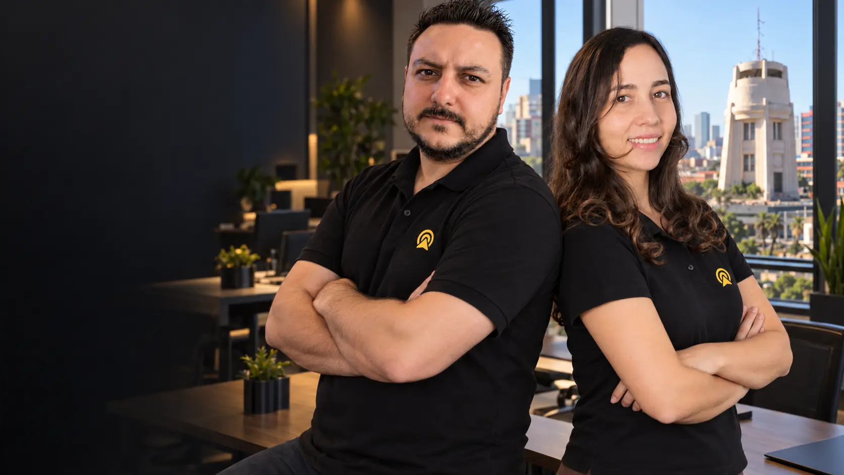Léo apoiado numa mesa e Lari de braços cruzados, costas com costas, olhando para a câmera no escritório da Arnoldi Digital