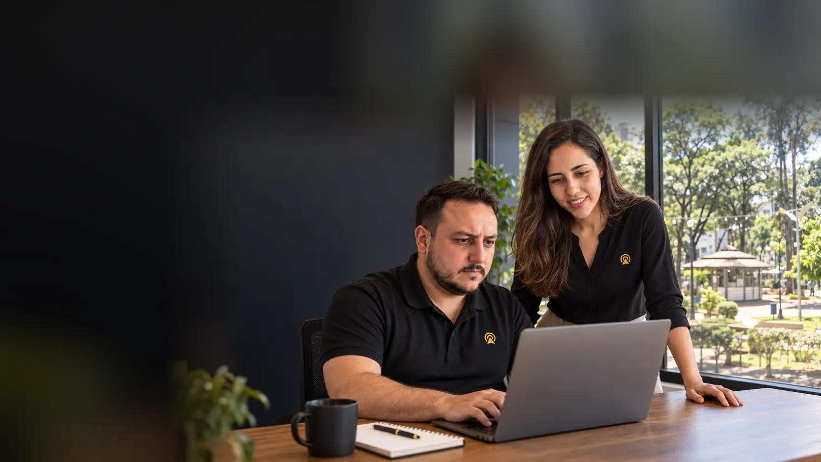 Léo e Lari, da Arnoldi Digital, analisando resultados de busca num notebook, com a Praça Comendador Müller de Americana desfocado ao fundo pela janela