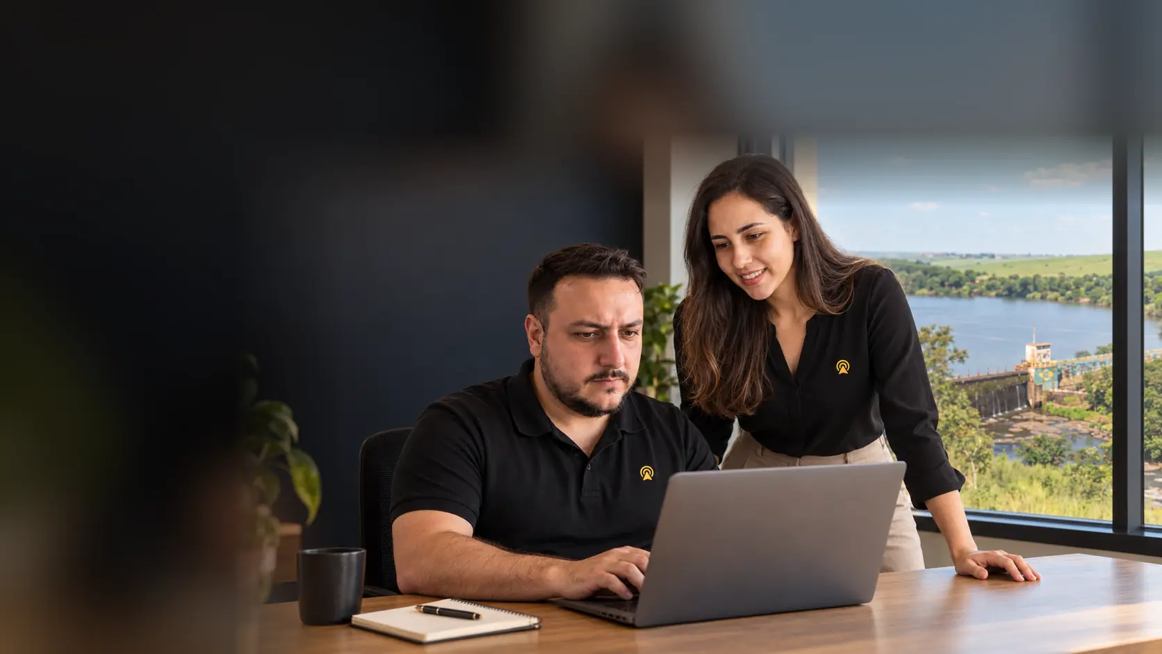 Léo e Lari, da Arnoldi Digital, analisando resultados de busca num notebook, com as ruínas da Usina Ester de Cosmópolis desfocado ao fundo pela janela