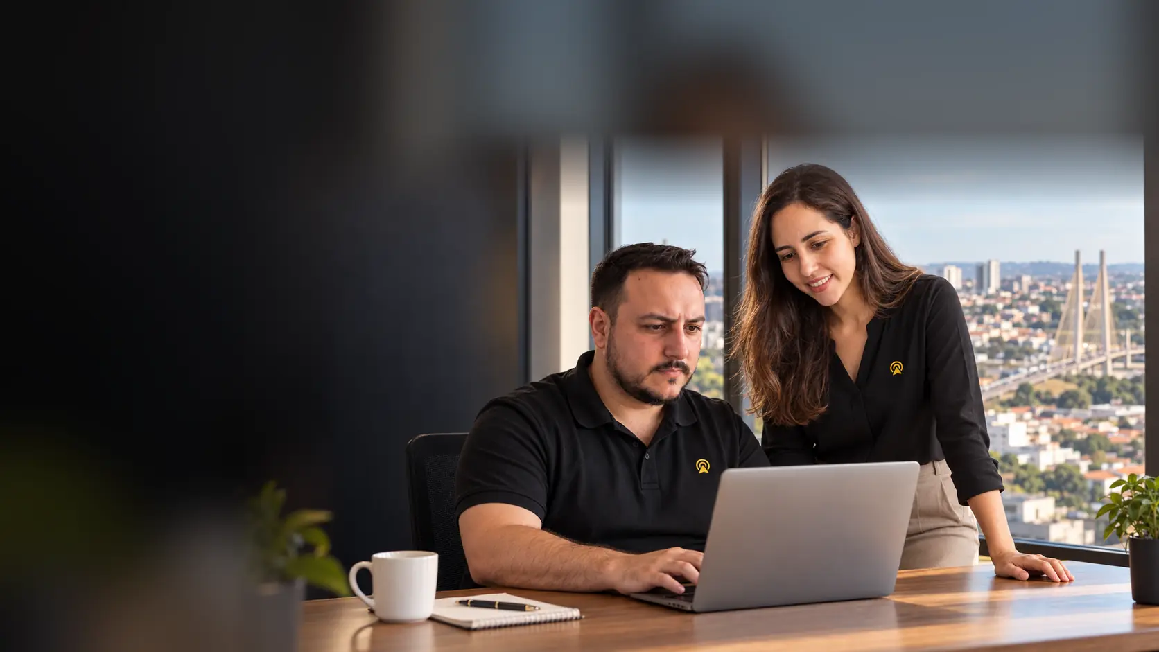 Léo e Lari, da Arnoldi Digital, analisando resultados de busca num notebook, com a Ponte Estaiada de Hortolândia desfocado ao fundo pela janela