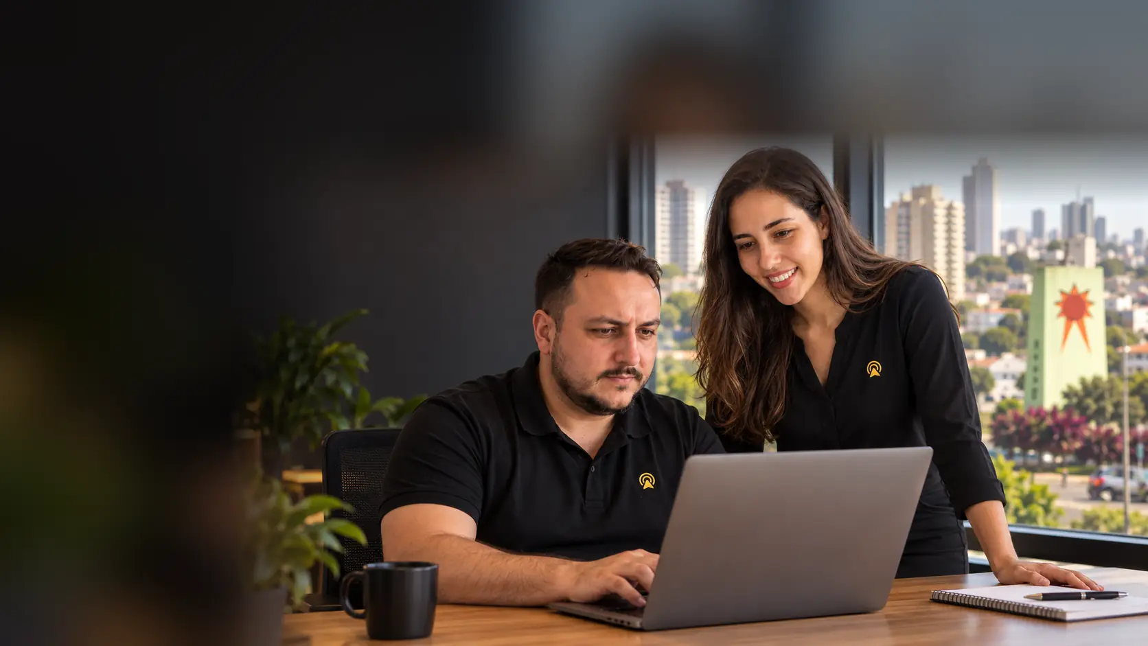 Léo e Lari, da Arnoldi Digital, analisando resultados de busca num notebook, com o Obelisco da Avenida Conceição de Indaiatuba desfocado ao fundo pela janela