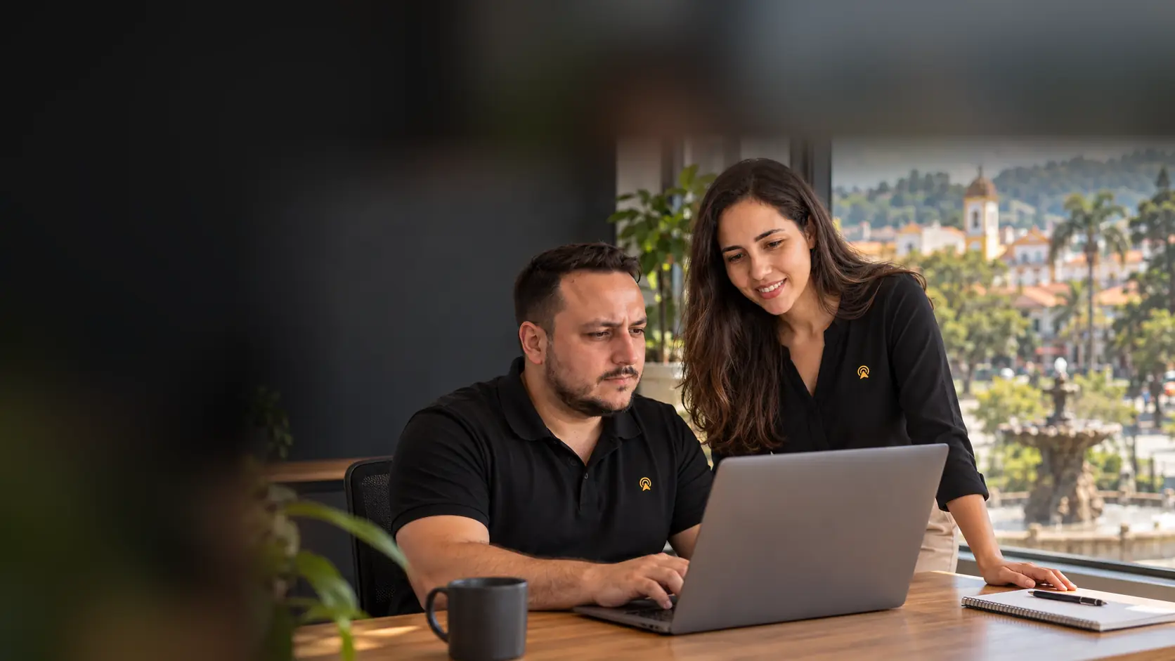 Léo e Lari, da Arnoldi Digital, analisando resultados de busca num notebook, com o chafariz histórico do centro de Itatiba desfocado ao fundo pela janela
