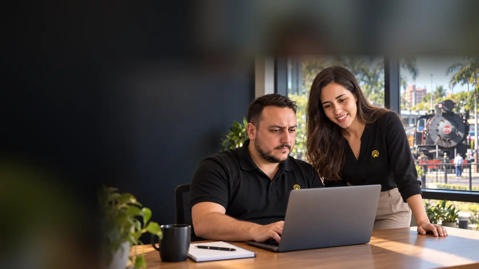 Léo e Lari, da Arnoldi Digital, analisando resultados de busca num notebook, com a Maria Fumaça de Jaguariúna desfocado ao fundo pela janela