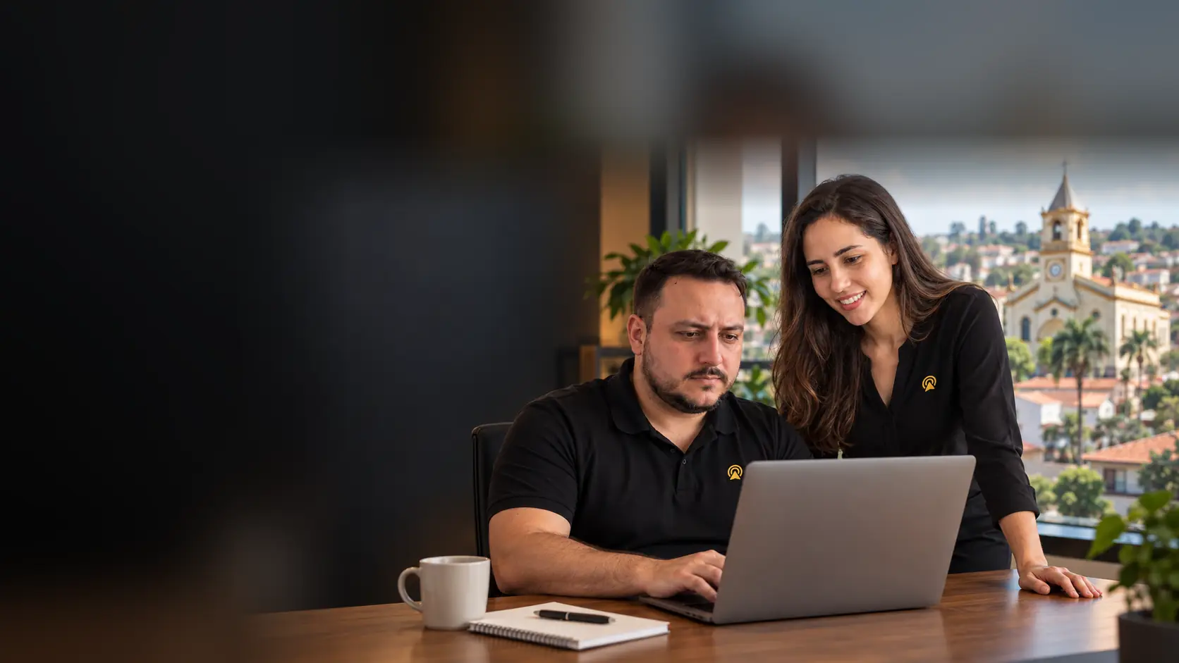 Léo e Lari, da Arnoldi Digital, analisando resultados de busca num notebook, com a Igreja de São Benedito de Monte Mor desfocado ao fundo pela janela
