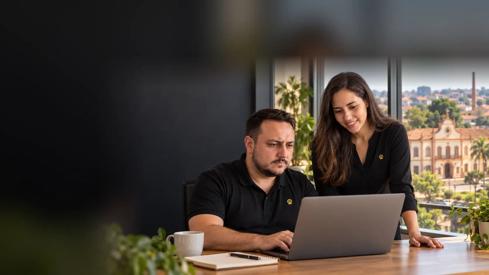 Léo e Lari, da Arnoldi Digital, analisando SEO num notebook, com o prédio histórico do Núcleo Colonial de Nova Odessa desfocado ao fundo pela janela