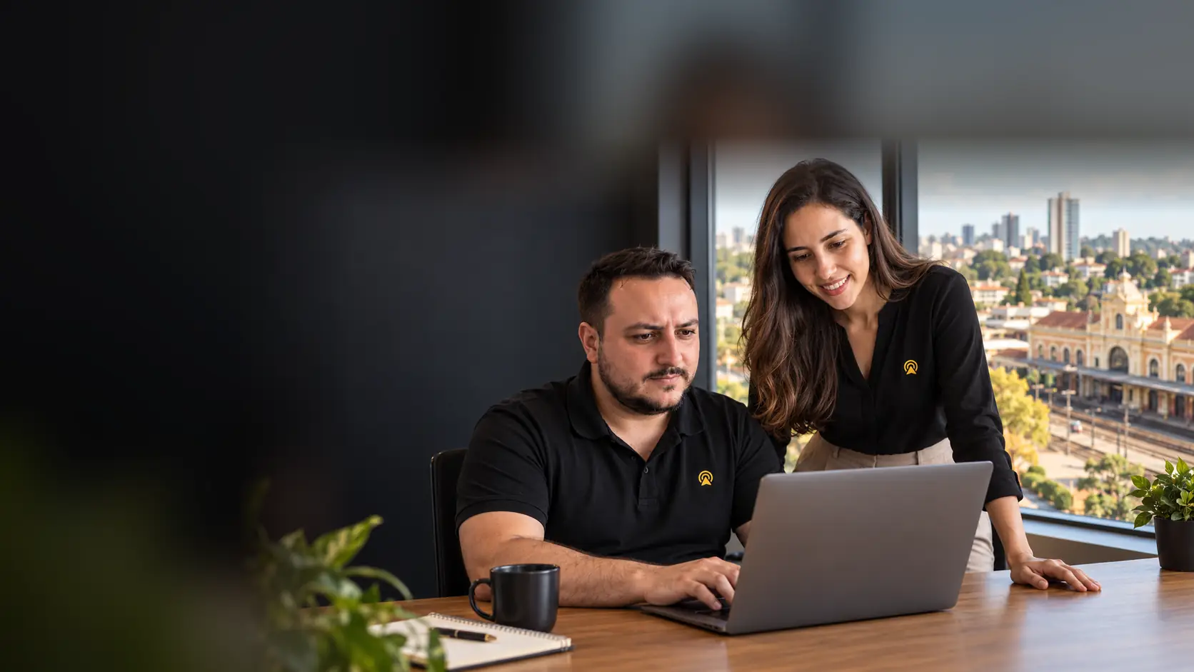 Léo e Lari, da Arnoldi Digital, analisando SEO num notebook, com a estação ferroviária histórica de Santa Bárbara d’Oeste desfocado ao fundo pela janela