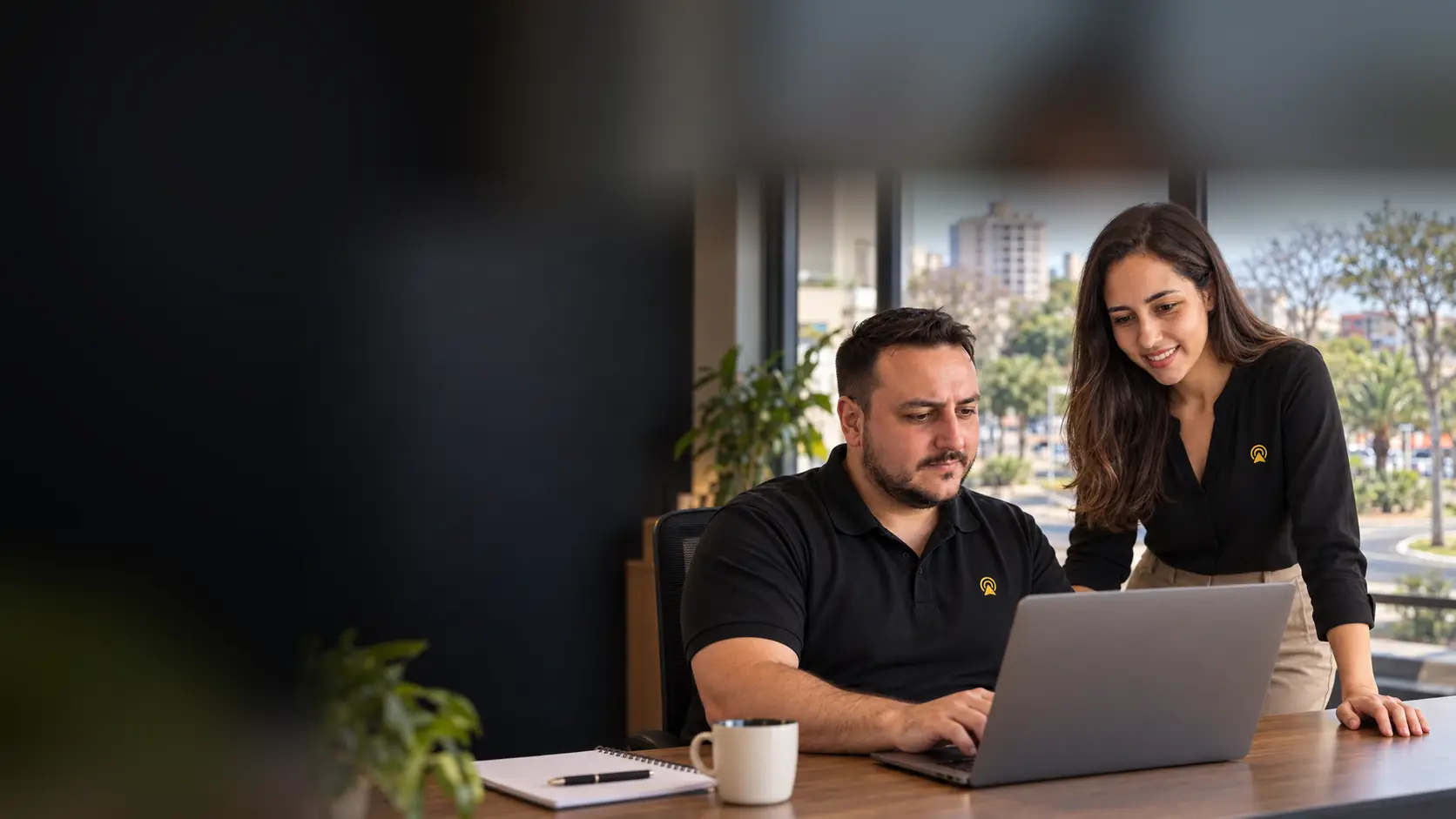 Léo e Lari, da Arnoldi Digital, analisando resultados de busca num notebook, com a Praça Brasil 500 Anos de Valinhos desfocado ao fundo pela janela