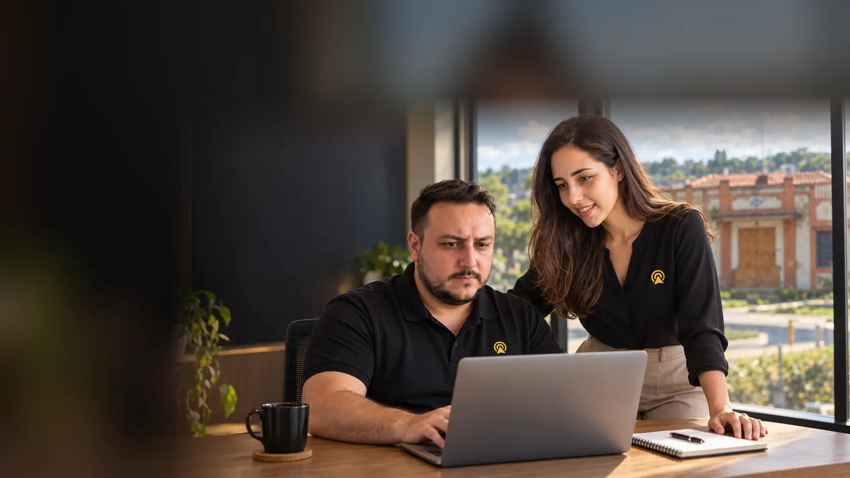 Léo e Lari, da Arnoldi Digital, analisando resultados de busca num notebook, com a estação ferroviária histórica de Vinhedo desfocado ao fundo pela janela