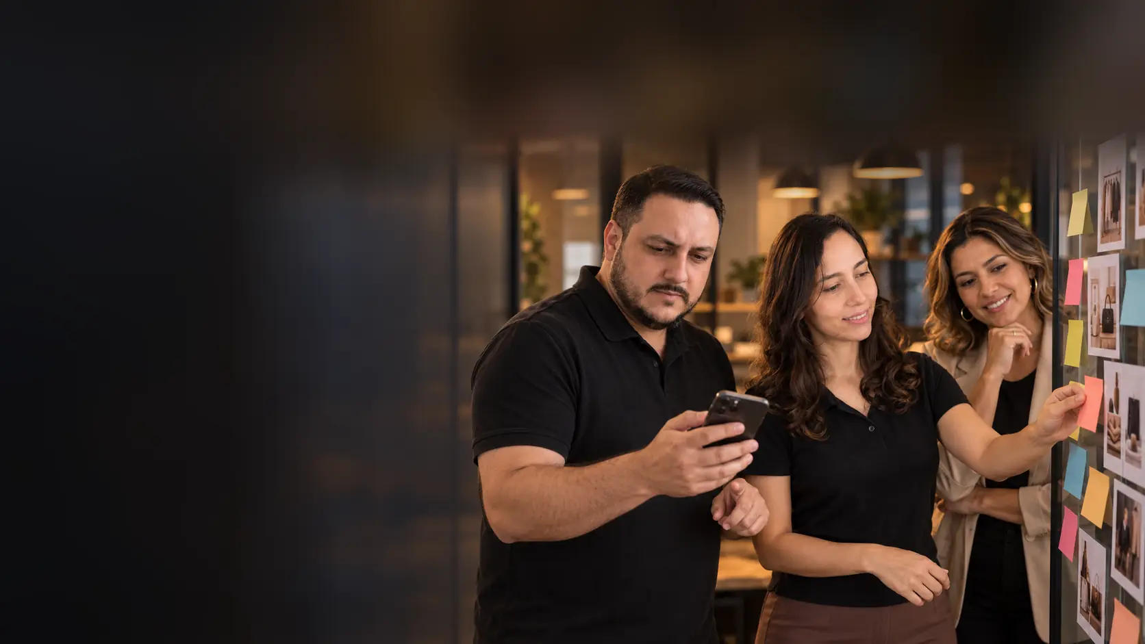 Léo olhando publicações no celular enquanto a Lari organiza um calendário de conteúdo com cartões coloridos e fotos de produtos em uma parede de vidro, ao lado de uma empresária