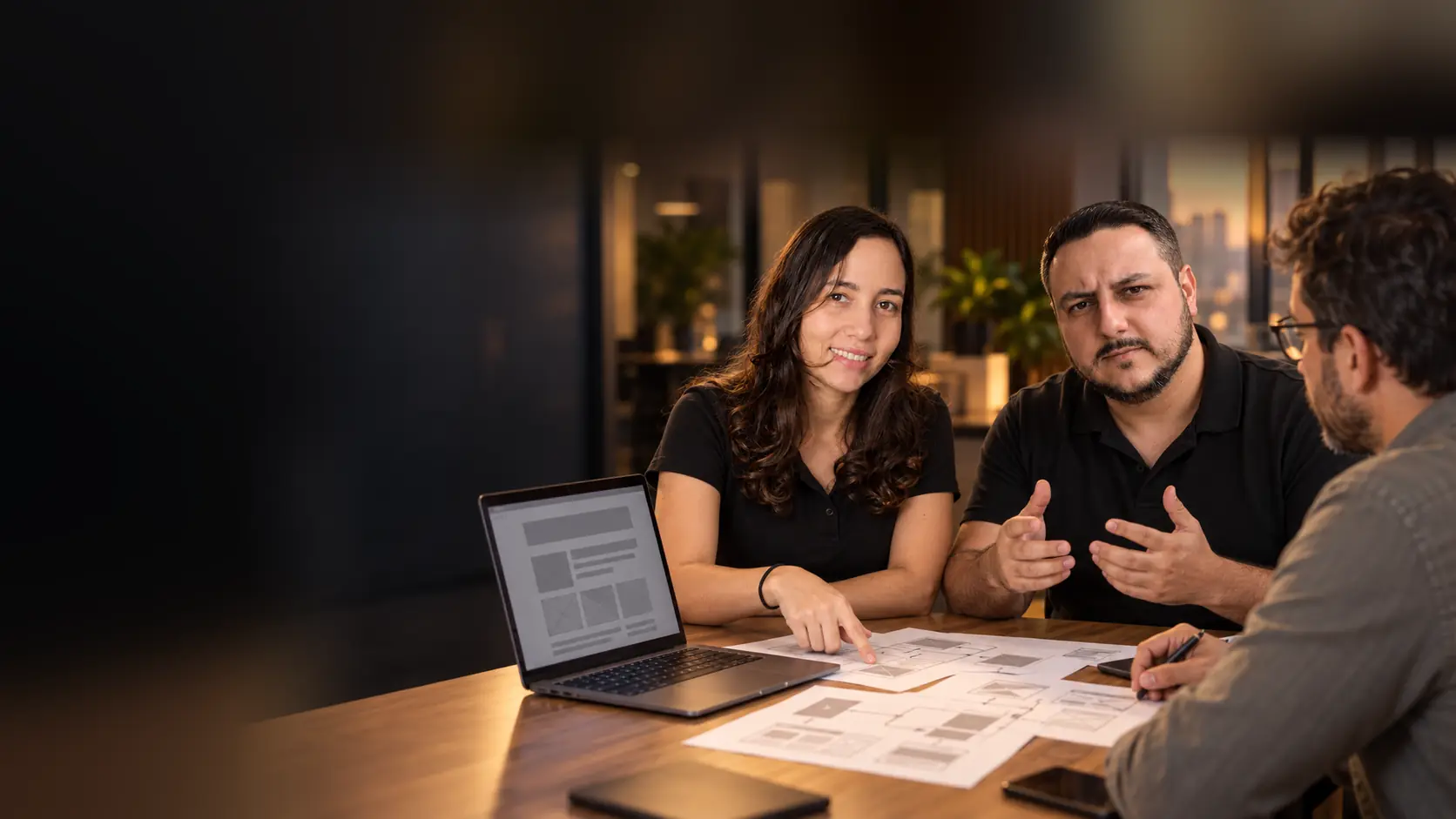 Léo e Lari em reunião com um empresário, com o rascunho de um site no notebook e esboços de páginas impressos sobre a mesa