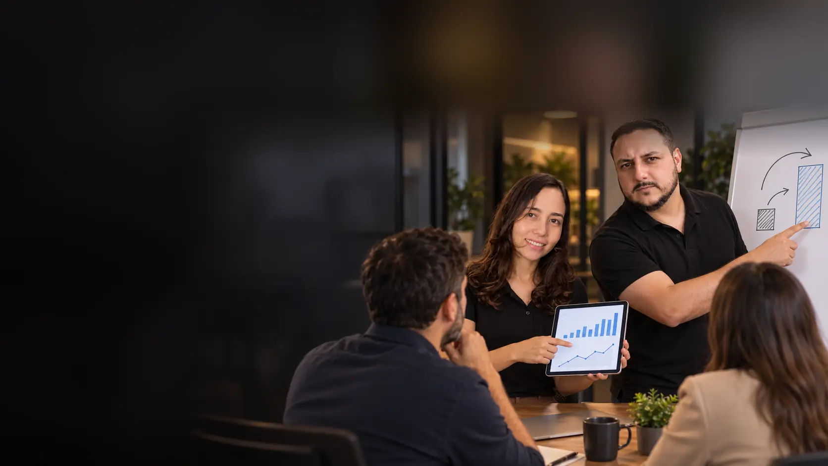 Lari mostrando a um casal de empresários um painel de anúncios no tablet enquanto o Léo aponta a divisão de um orçamento desenhada no quadro branco