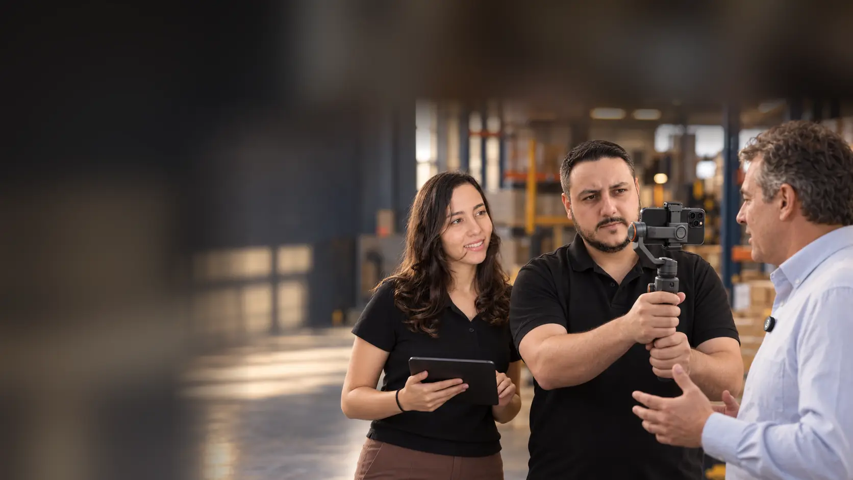 Léo gravando com um celular no gimbal de mão um empresário com microfone de lapela sem fio, enquanto a Lari acompanha o roteiro no tablet, dentro de um galpão